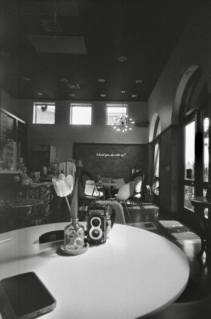 Rolleiflex twin-lens camera and a tulip in a glass bottle on a café table, with a sputnik chandelier and a “Would you sit with us?” neon sign, Magnolia Coffee Bar (former), Art Village on Main, McAllen, Texas — Arista 400 35mm, RETO Ultra Wide & Slim