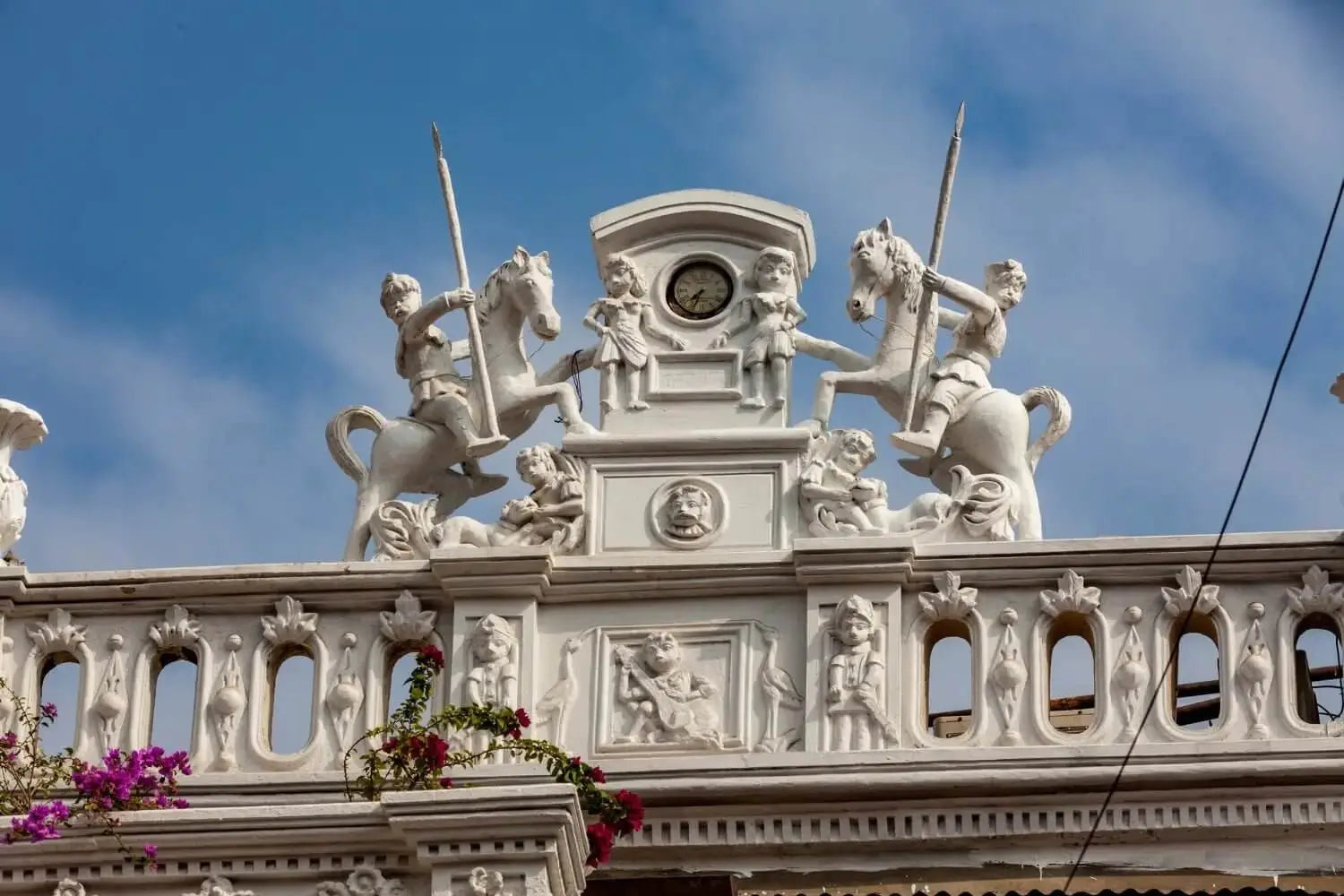 White ornate clock tower with sculpted figures of knights on horseback and children, set against a blue sky.