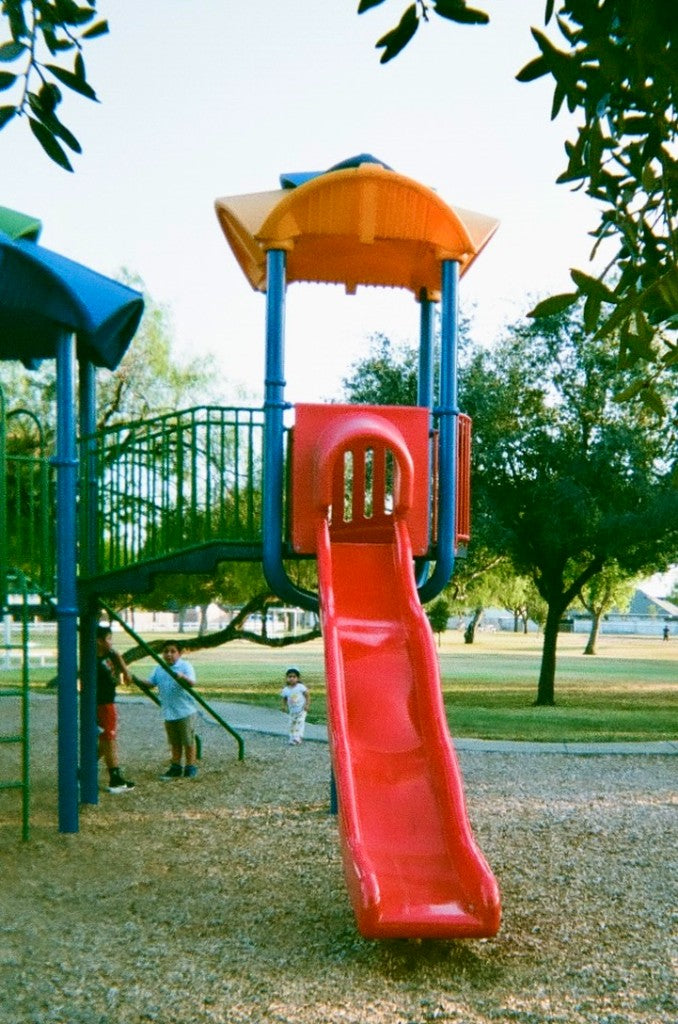 Red playground slide with kids in a sunlit park, shot on Fuji Quicksnap 400 Iso Color C41 Disposable Film Camera, developed in Kodak C-41