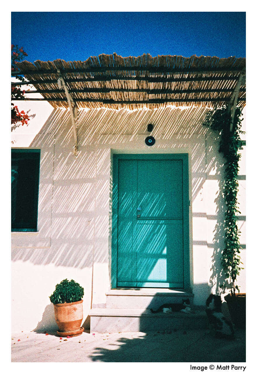 A vibrant teal-colored door with a simple rectangular design and vertical paneling, set within a white-walled entrance under a thatched roof.