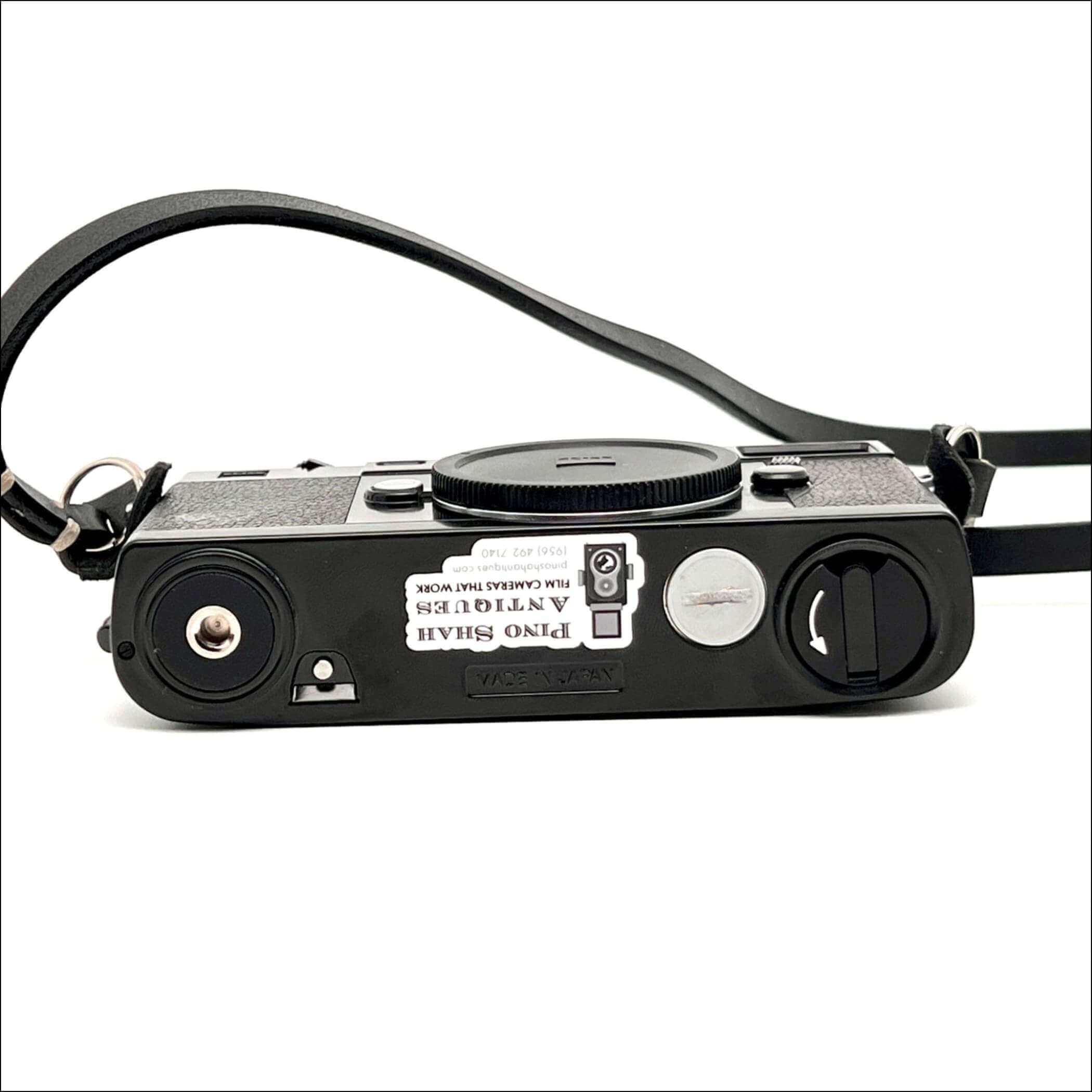 Black textured body with a silver shutter button and black leather strap, featuring a sticker that reads ’pino shah antiques film cameras that work.’