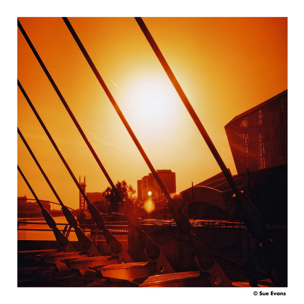A modern bridge with sleek, dark metal cables stretching diagonally across a warm orange sunset sky.