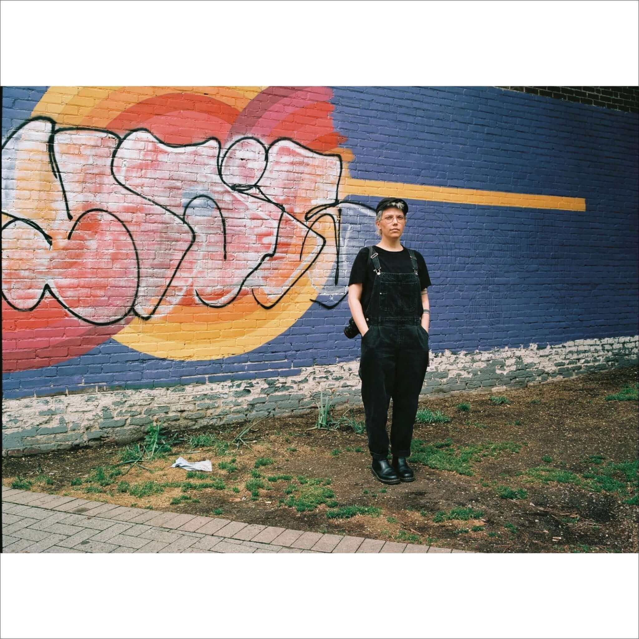 A person wearing black denim overalls with a short-sleeved shirt and dark shoes stands in front of a colorful graffiti mural.