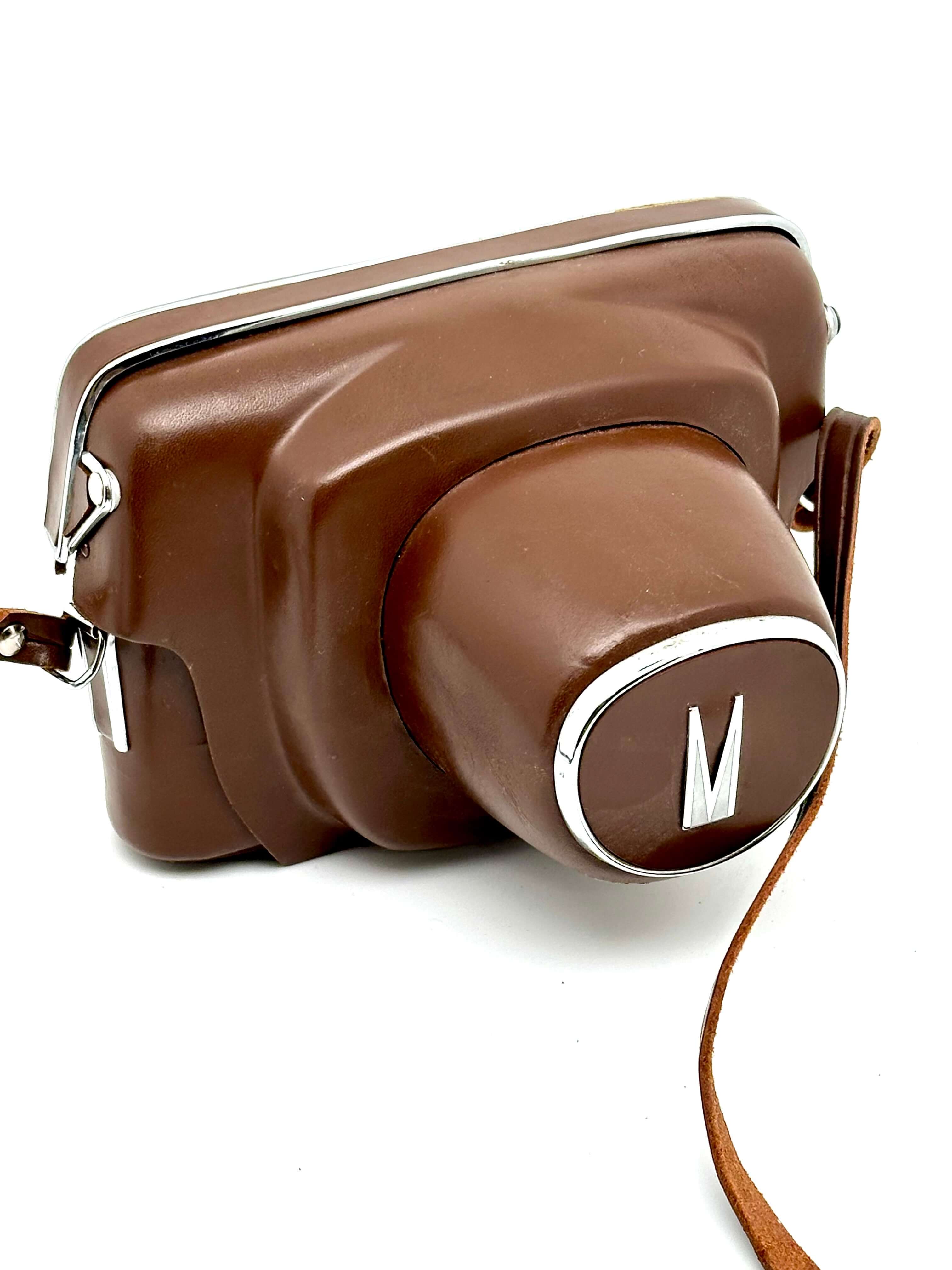 Brown leather camera case with a silver ’m’ logo on the lens cover and matching strap.