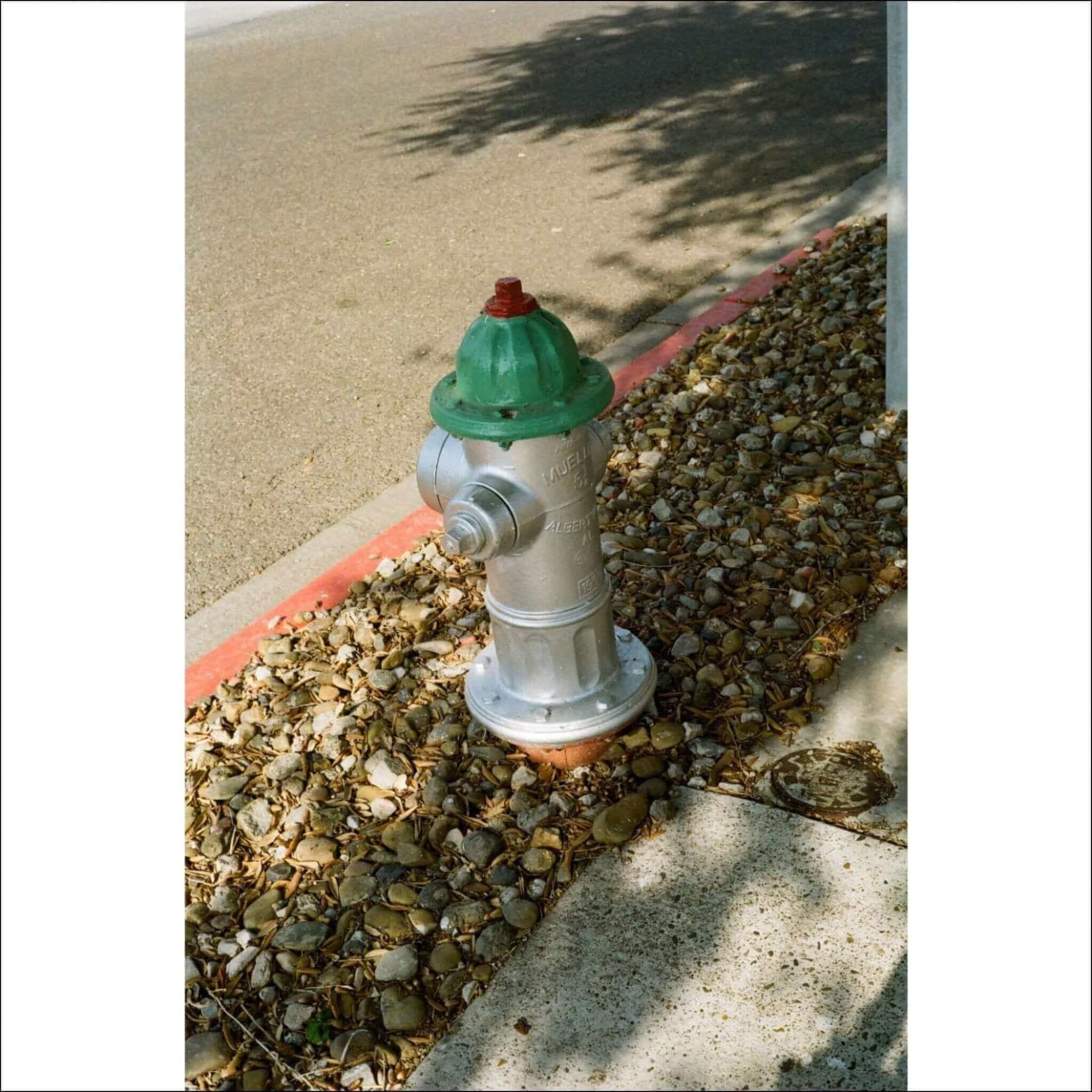 A silver fire hydrant with a green top and red cap, set in a gravel bed beside a sidewalk.