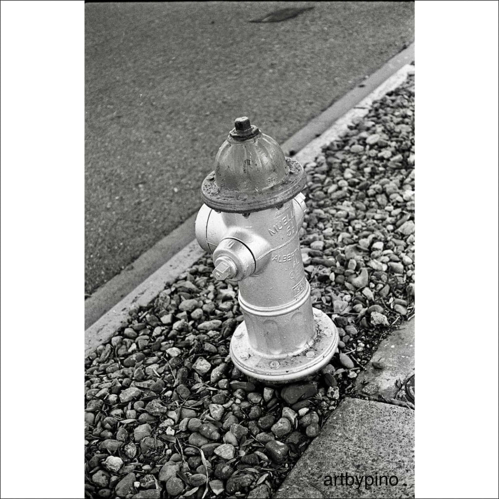 Metallic fire hydrant with a domed top and bolted cap, standing on gravel beside a curb.