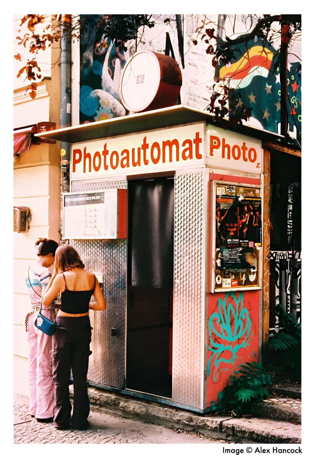 A vintage-style photo booth with a corrugated metal exterior, red and white signage reading ’photoautomat photo,’ and colorful graffiti on its side panels.