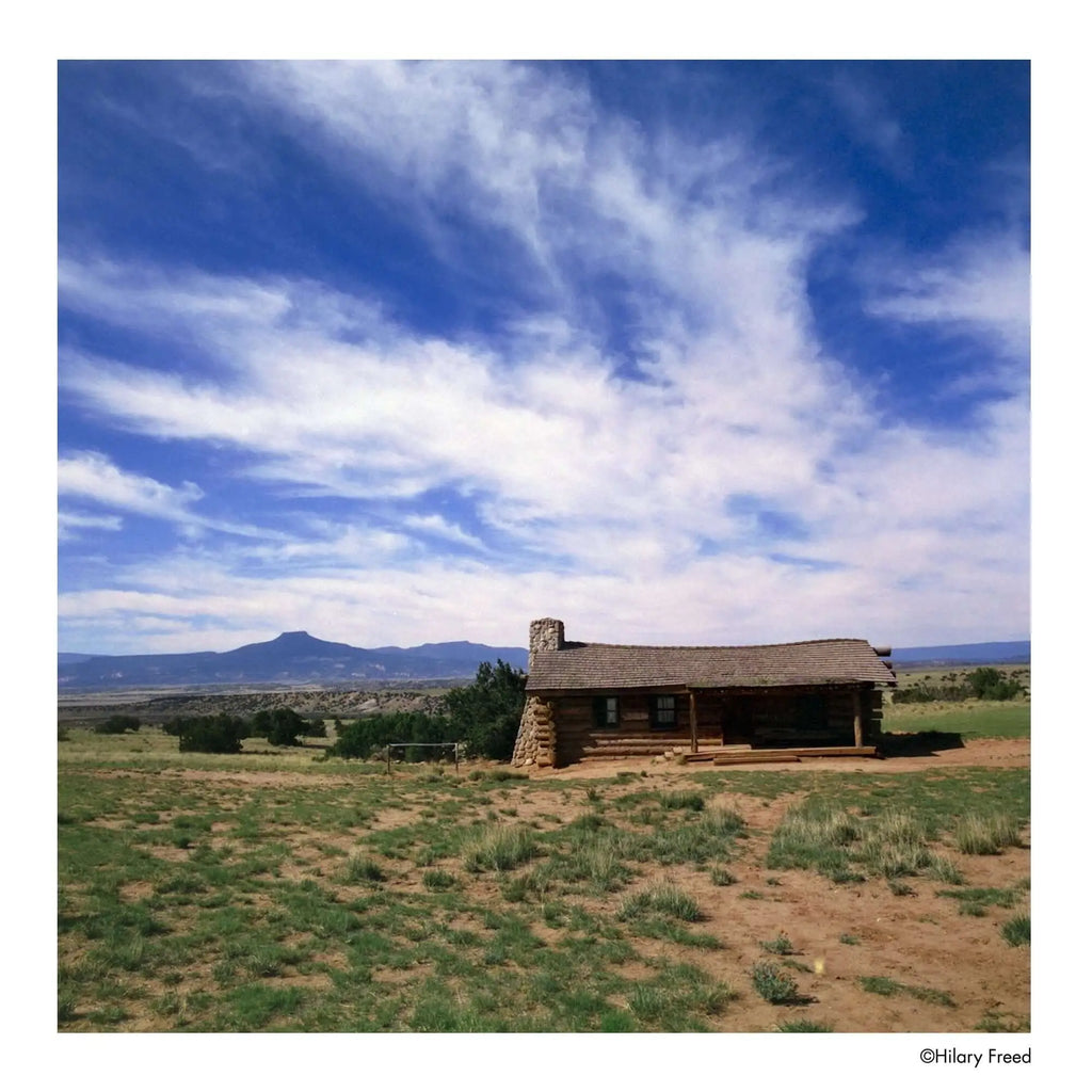 A rustic log cabin with a stone chimney and wooden roof, set against an expansive blue sky dotted with wispy clouds.
