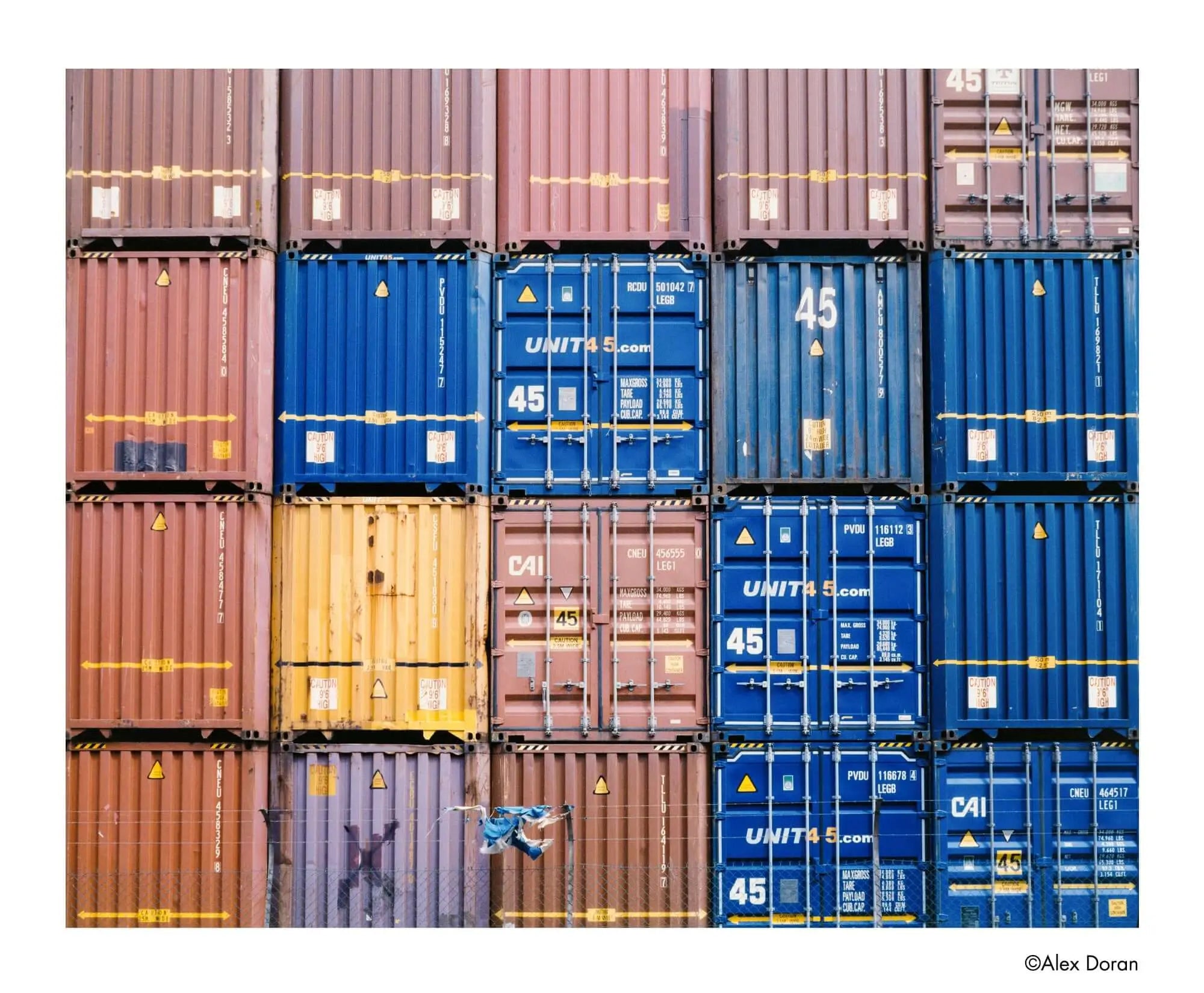 Stacked shipping containers in muted reds, blues, yellows, and purples with yellow markings and ’unit4.com’ branding.
