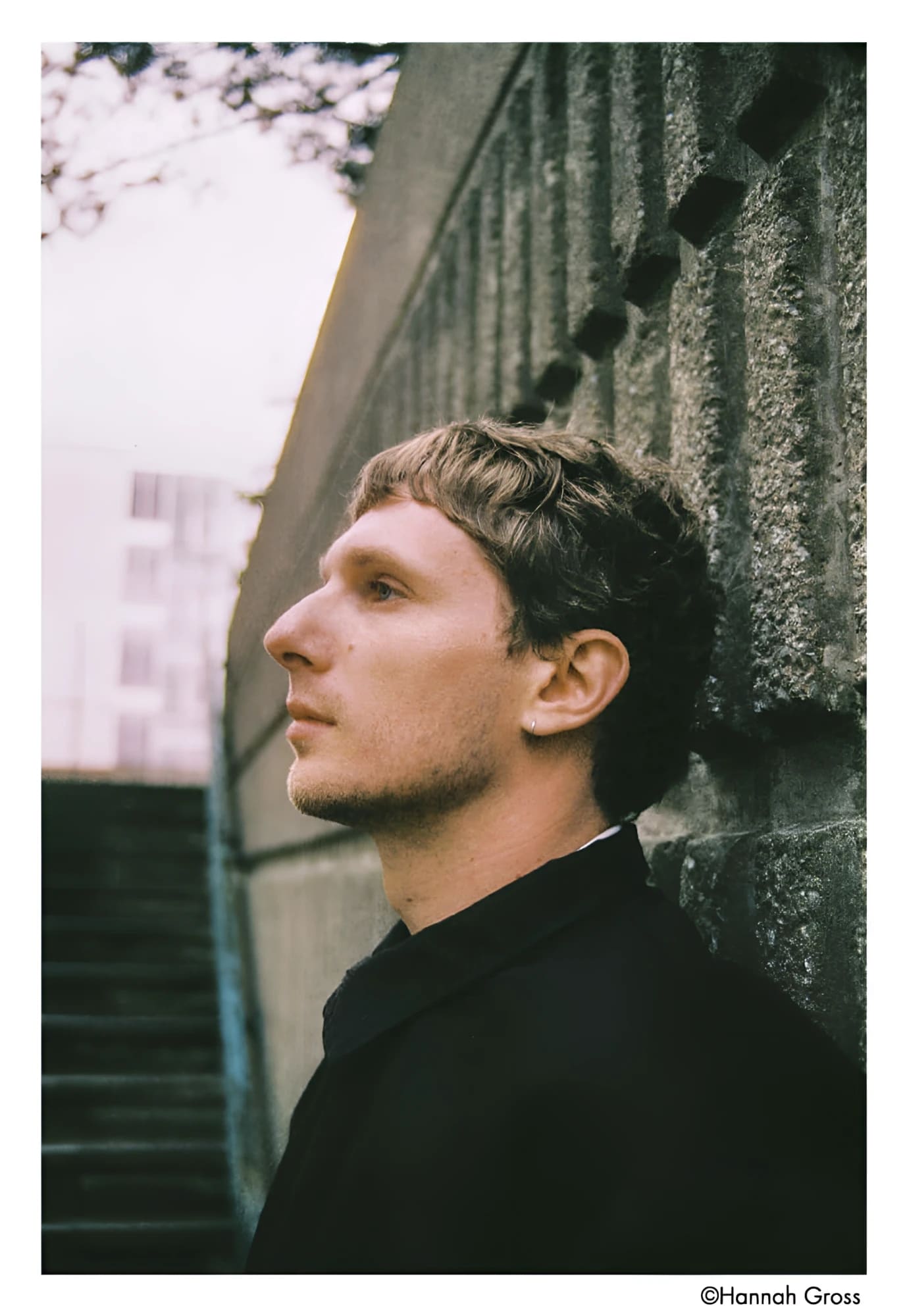 A man with short brown hair and a light stubble, wearing a black coat.