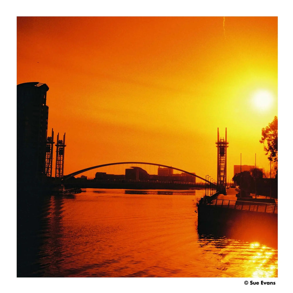 Silhouetted arched bridge spanning a river under a vibrant orange sunset sky.