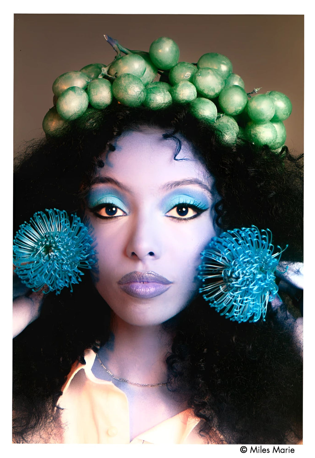 A woman wears a green grape crown and blue dandelion-like earrings.
