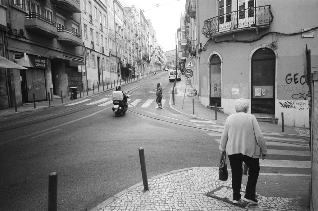 An elderly woman in a light-colored coat and dark pants walks across a cobblestone street, carrying a dark handbag.