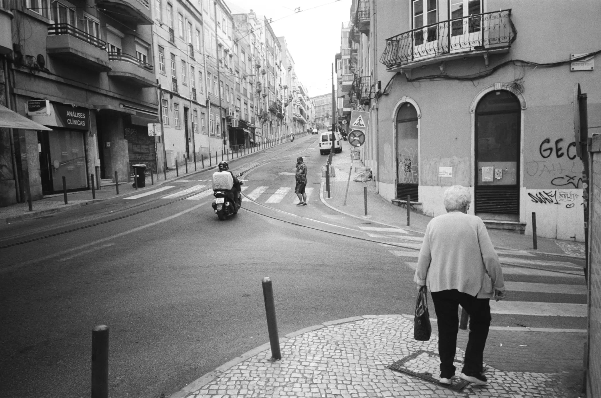 An elderly woman in a light-colored coat and dark pants walks across a cobblestone street, carrying a dark handbag.