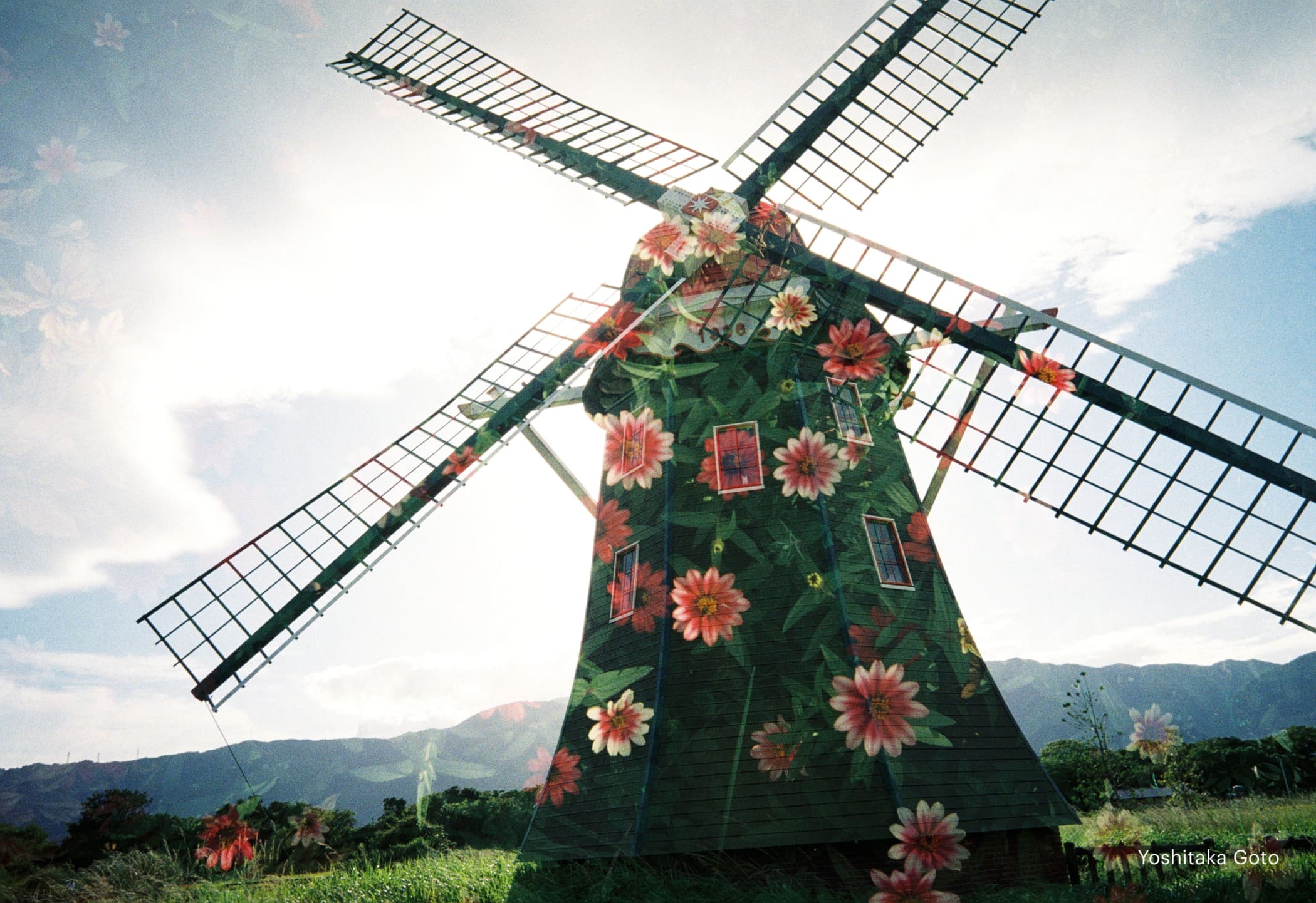 A green windmill adorned with vibrant pink and orange flowers, featuring a painted design on its wooden structure.