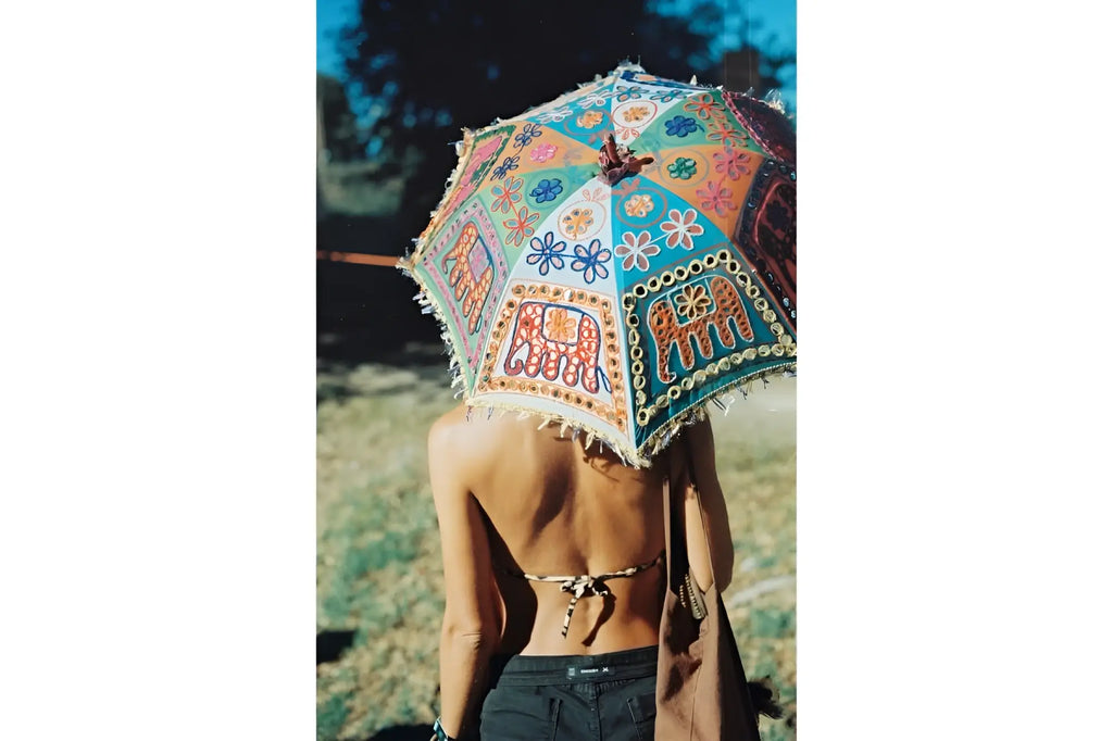 A vibrant, hand-painted indian-style umbrella with intricate floral and elephant motifs in turquoise, orange, red, and blue hues, adorned with decorative fringe along its edges.