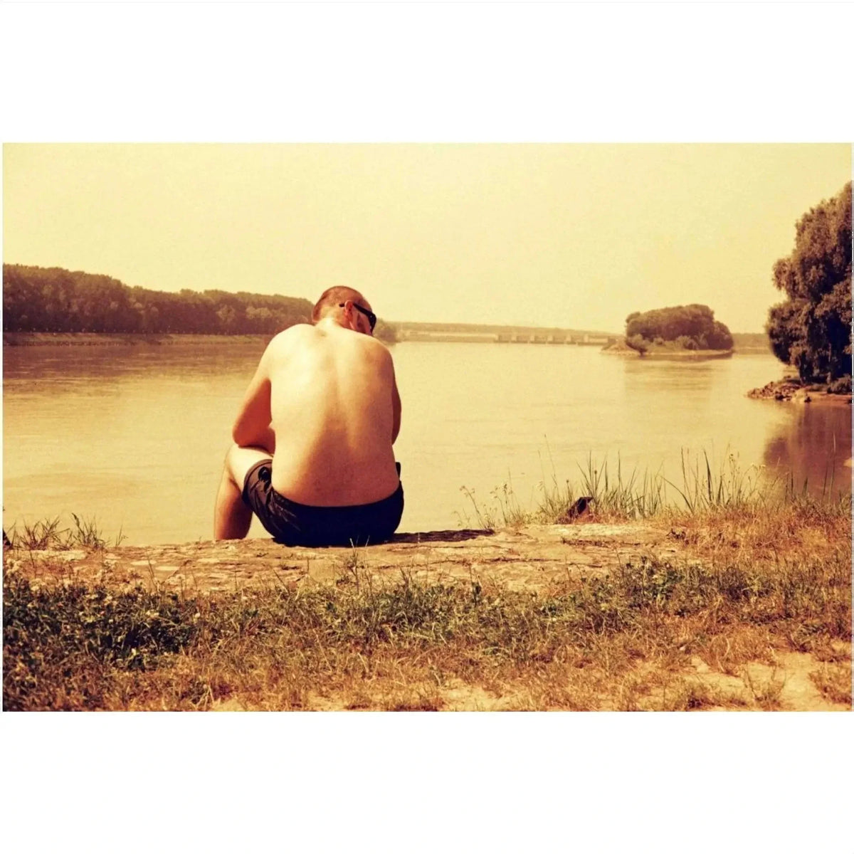 A man wearing black shorts sits on a grassy riverbank, facing away from the camera toward calm waters under a warm-toned sky.