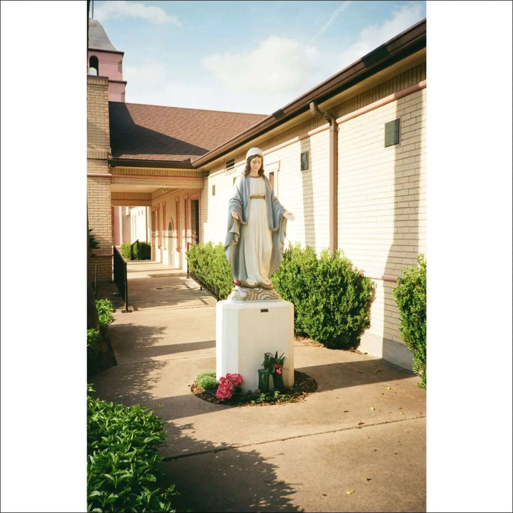 A serene white statue of the virgin mary, dressed in flowing robes with a blue sash and standing on an ornate pedestal.