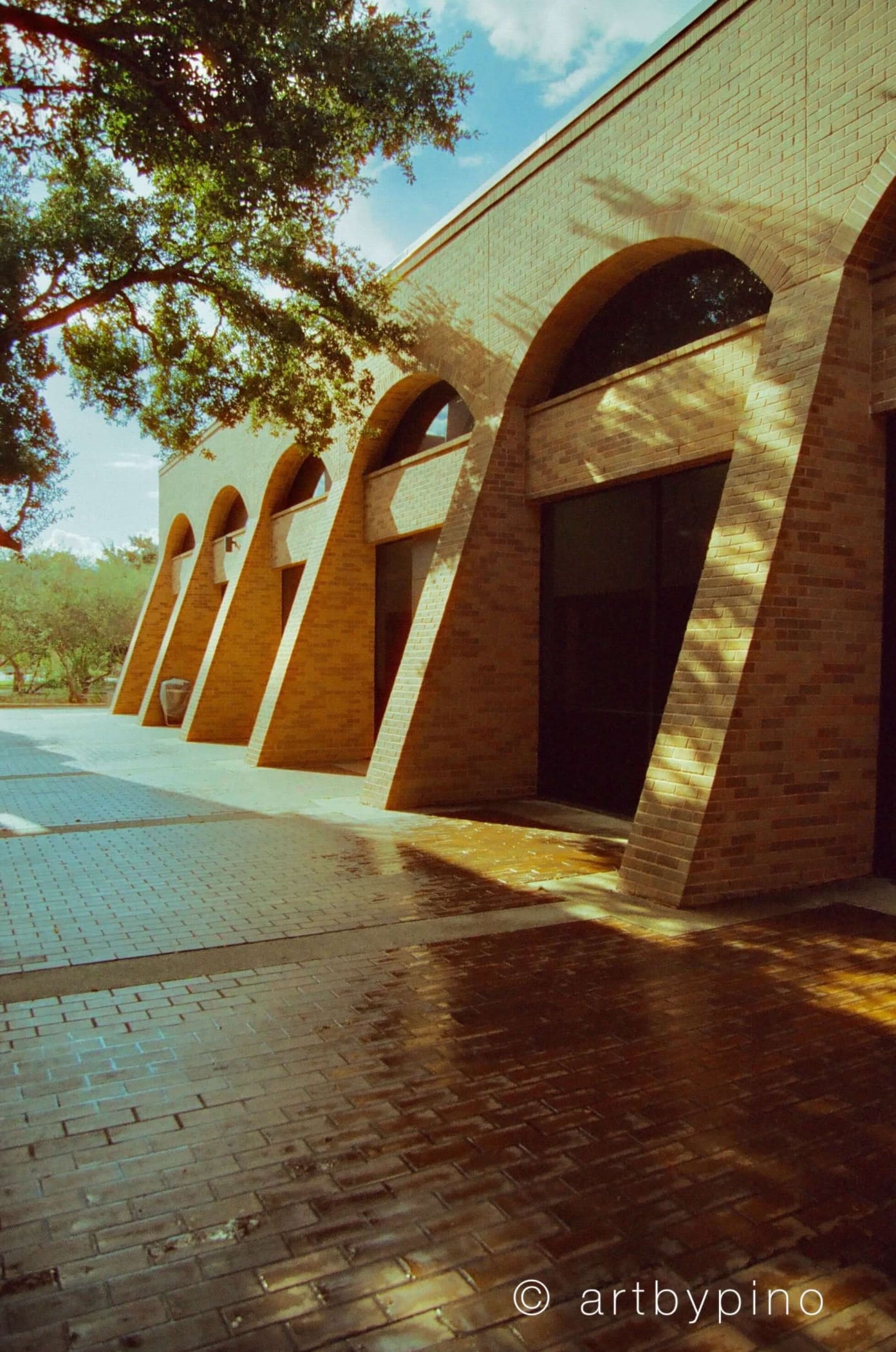 The building features a series of warm-toned brick arches with dark glass doors, set against a backdrop of trees and blue sky.