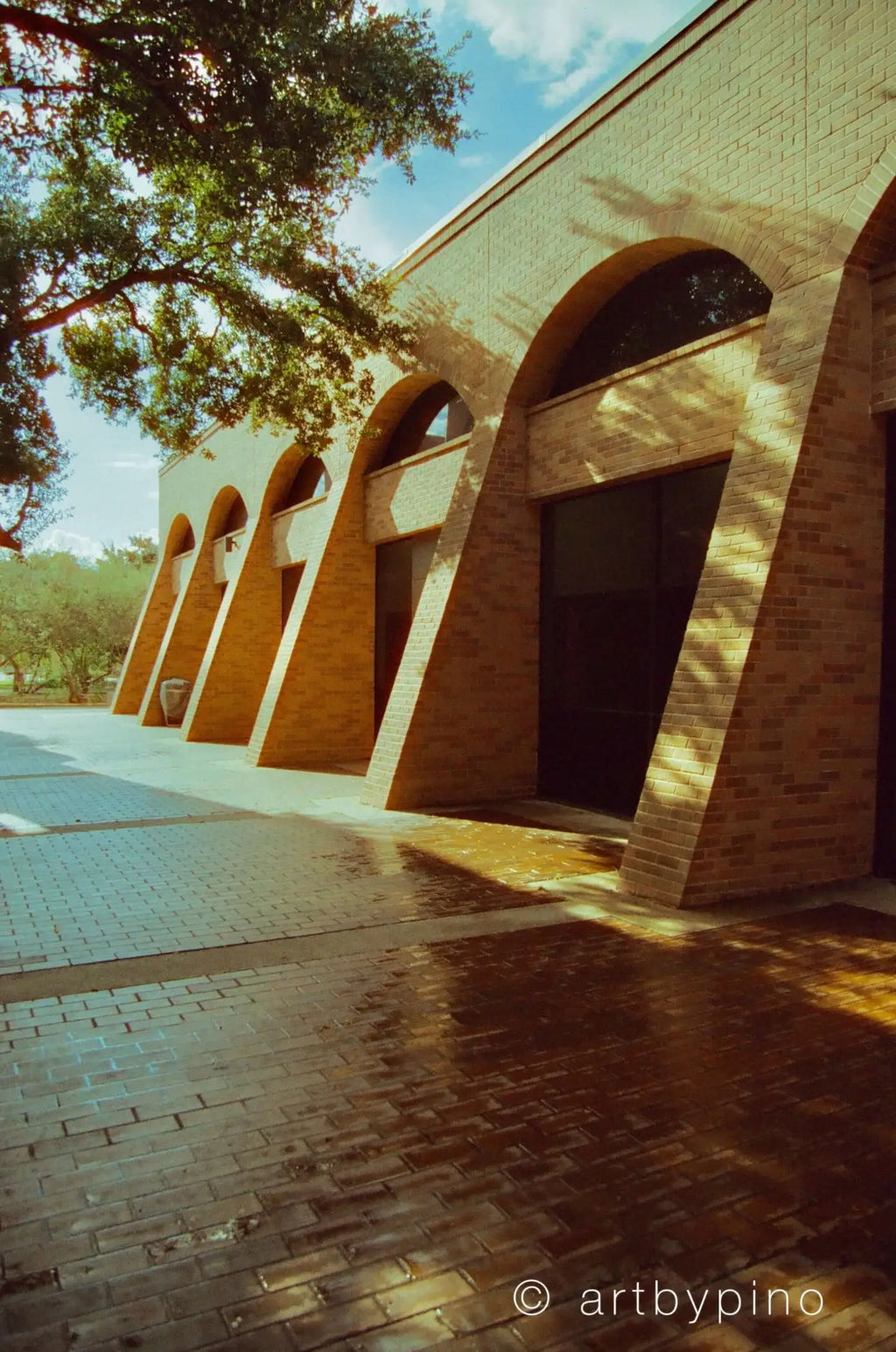 The building features a series of warm-toned brick arches with dark glass doors, set against a backdrop of trees and blue sky.