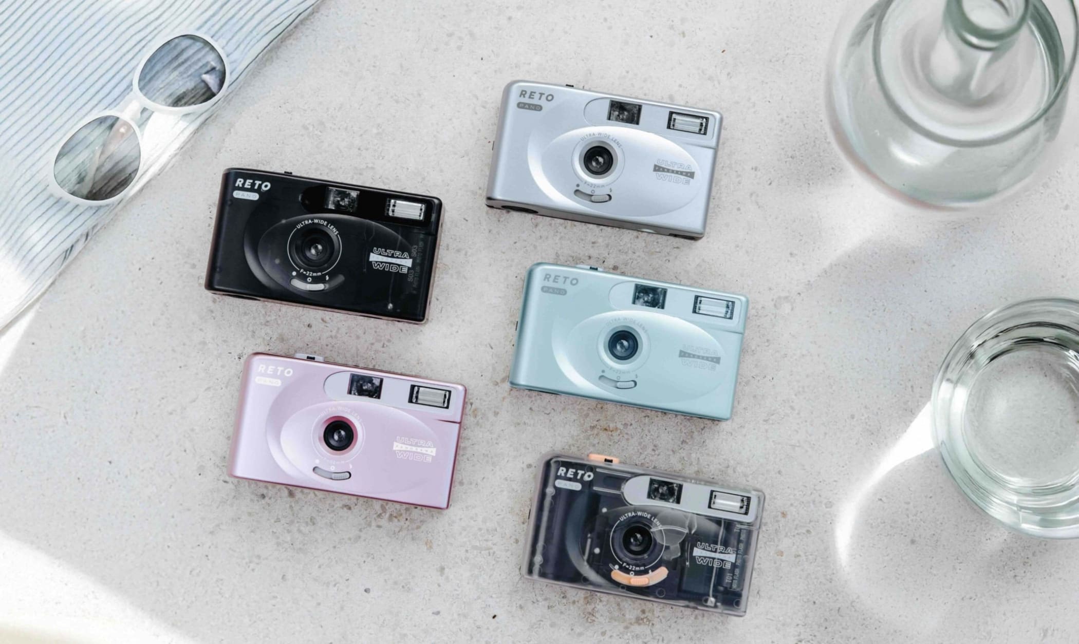 Five compact reto brand instant cameras in black, silver, light blue, pink and transparent with orange accents.