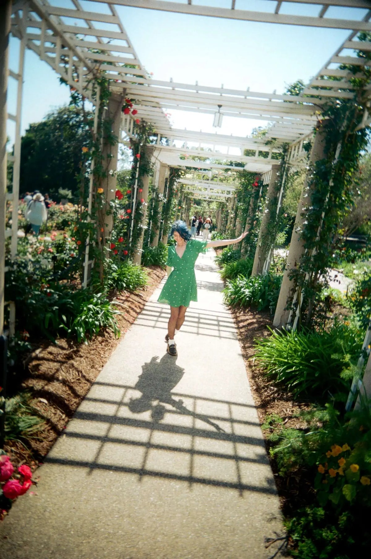 A woman in a vibrant green, short-sleeved dress with small white polka dots walks under a white wooden arbor.